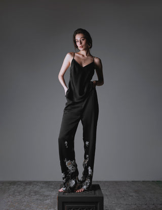Woman wearing a black silk set composed by long pants and a v neck top with spaghetti straps and with floral patterns on a gray background