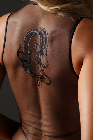 A close-up of a woman's bare back wearing sheer black mesh lingerie, featuring a large black ram tattoo design on the upper back.