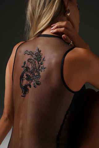 A woman shown from the back wearing a black sheer mesh camisole, displaying a large black floral vine tattoo on her upper back. She is wearing gold hoop earrings and a gold ring, set against a dark studio background.