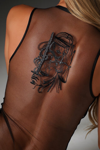 A close-up of a woman's bare back showing a large black graphic tattoo of a woman holding a sword, wearing a sheer black mesh lingerie bodysuit.
