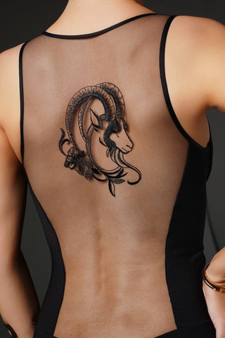 A black temporary tattoo of a ram's head with floral details, applied on the upper back of a model wearing a black strappy tank top.