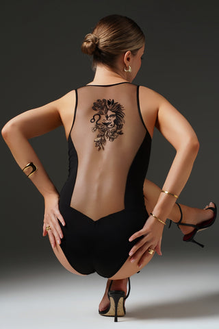 A woman photographed from behind wearing a black one-piece bodysuit with a sheer back panel and a large black floral lion-back graphic. She is posed in a crouching position against a dark gray studio background.