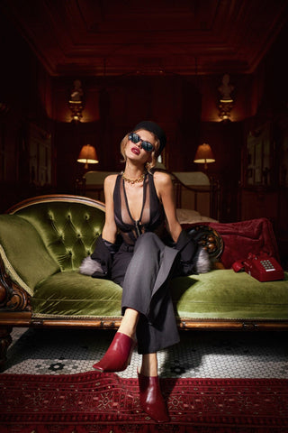 Woman in a stylish outfit sitting on a green couch in a dimly lit room. Lingerie Devil Bodysuit