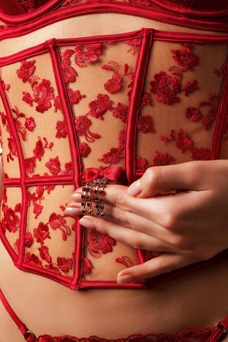 Close-up of a person wearing a red lace and mesh corset-style lingerie piece, with a hand resting on the front over a decorative ring.