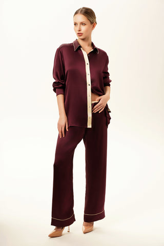 Woman wearing a silk satin burgundy pajama set with a beige edge contract and button-down shirt. Long silk pants. White background