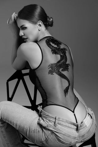 Woman sitting on a chair wearing a sheer top with a dragon design, gray background